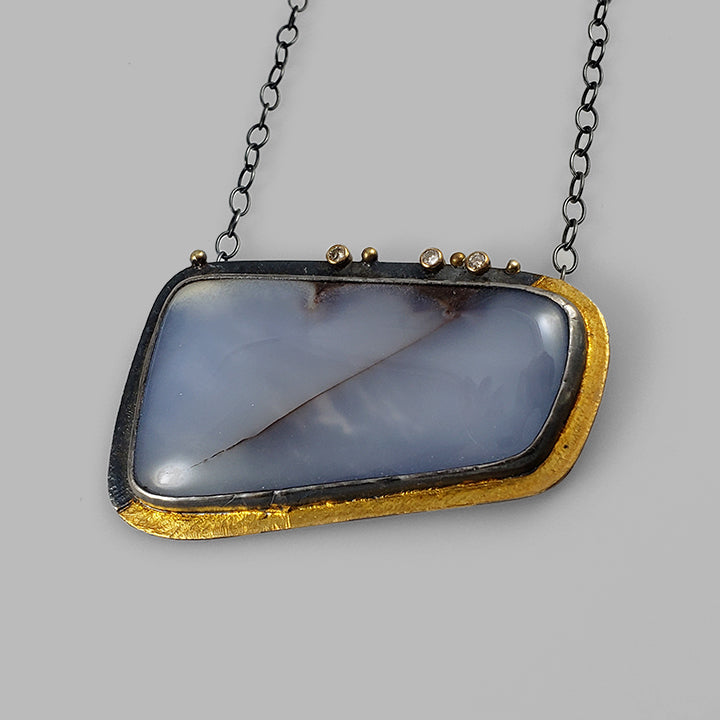 large periwinkle gemstone set in oxidized silver on textured plate in silver and gold with diamonds